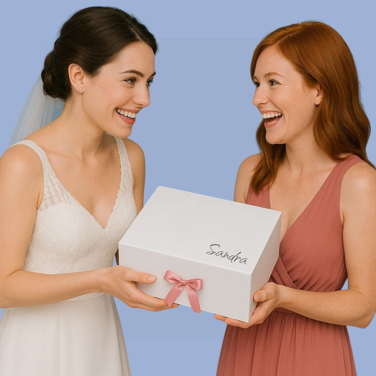 Braut schenkt ihrer Trauzeugin eine weiße Box mit rosa Schleife. Beide lachen sich an, vor blauem Hintergrund.