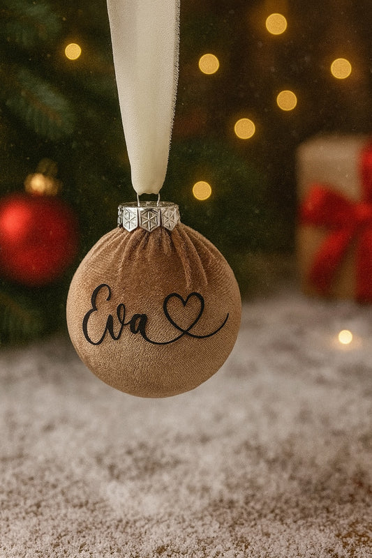 Personalisierte Weihnachtskugel in Braun, Ø 8 cm, beflockt mit Samt-Oberfläche und silbernem Kranz, verziert mit dem Namen ‚Eva‘ und Herzmotiv. Aufgehängt am weißen Chiffonband, vor weihnachtlichem Hintergrund mit Baum, Lichtern und Geschenken.