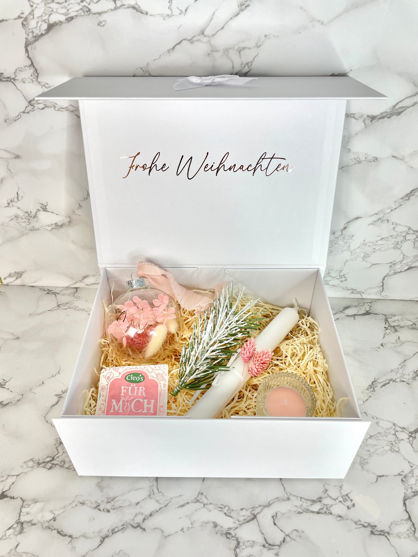 Weiße Weihnachtsgeschenkbox mit Aufdruck ‚Frohe Weihnachten‘, gefüllt mit personalisierbarer Weihnachtskugel mit rosa Trockenblumen, weißer Stabkerze mit Deko, Kunst-Tannenzweig, Teelicht im Glas und Kräutertee, dekoriert mit Holzwolle.