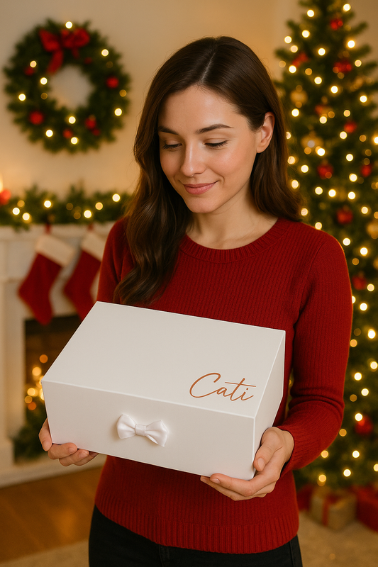 Frau im roten Pullover hält eine weiße Geschenkbox mit Personalisierung ‚Cati‘ in den Händen, vor weihnachtlich geschmücktem Wohnzimmer mit Lichterkette, Weihnachtsbaum und Kranz.