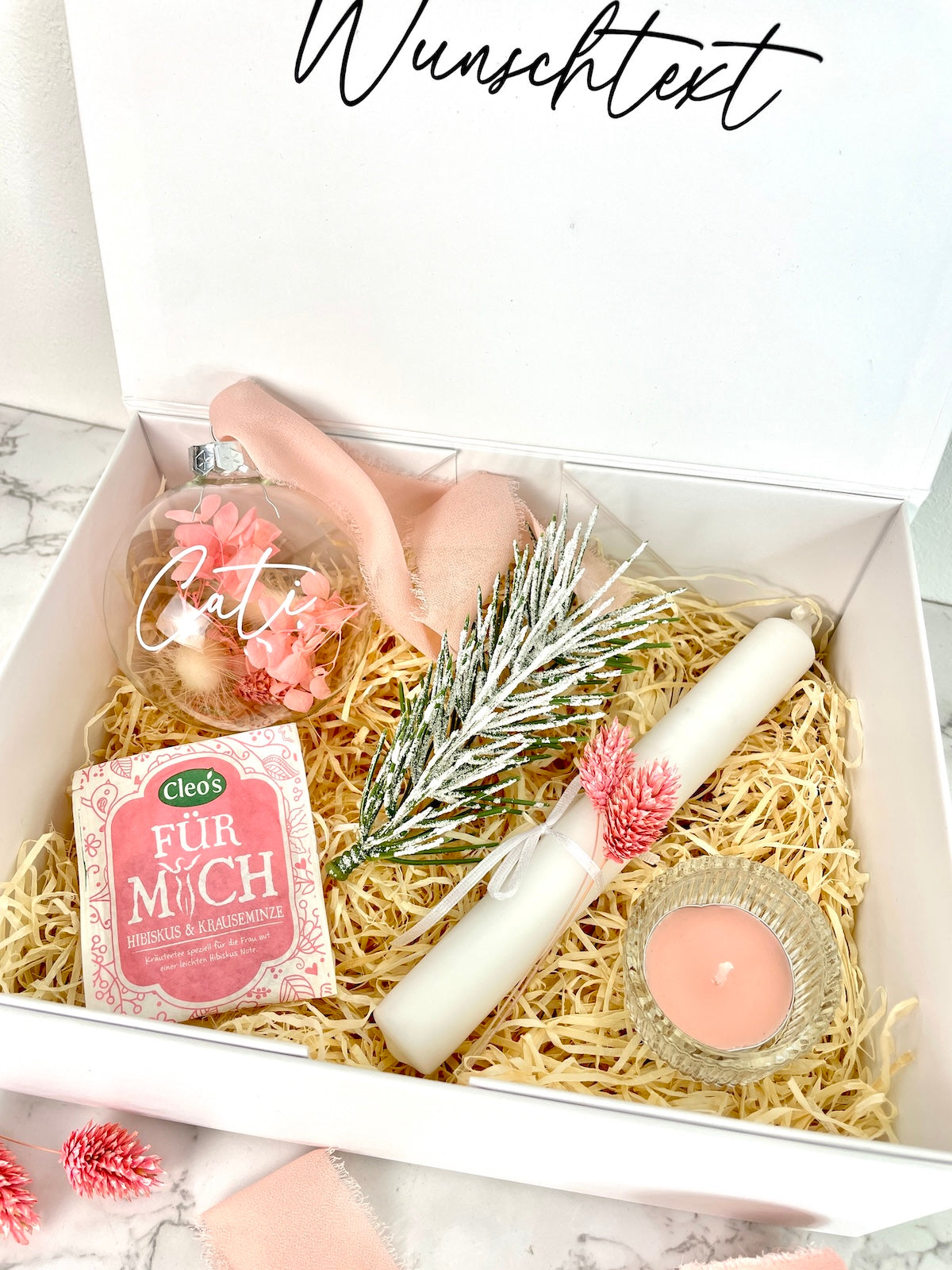 Personalisierte weiße Geschenkbox mit Wunschtext, daneben der Inhalt: Glas-Weihnachtskugel mit rosa Trockenblumen und Chiffonband, weißer Kunst-Tannenzweig mit Schnee-Effekt, weiße Stabkerze mit rosa Trockenblumen, Kräutertee von Cleo’s und rosa Duft-Teelicht im Glas.