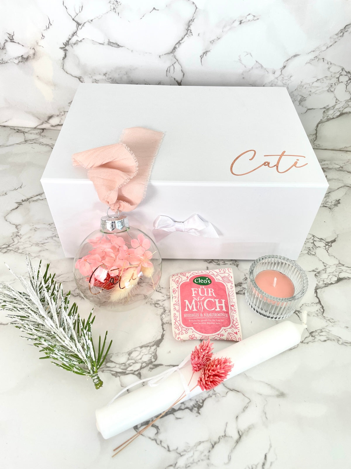 Personalisierte weiße Geschenkbox mit Namensaufdruck ‚Cati‘, daneben der Inhalt: Glas-Weihnachtskugel mit rosa Trockenblumen und Chiffonband, weißer Kunst-Tannenzweig mit Schnee-Effekt, weiße Stabkerze mit rosa Trockenblumen, Kräutertee von Cleo’s und rosa Duft-Teelicht im Glas.