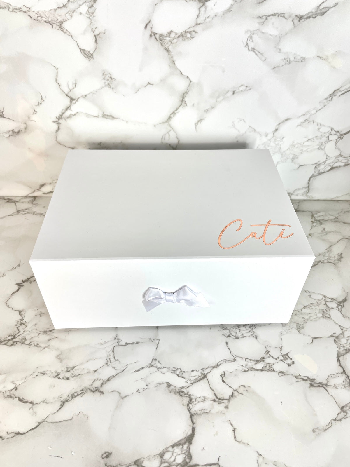Weiße, stabile Geschenkbox mit weißer Schleife und personalisiertem Namensaufdruck ‚Cati‘ in Roségold, auf weißem Marmortisch dekoriert.