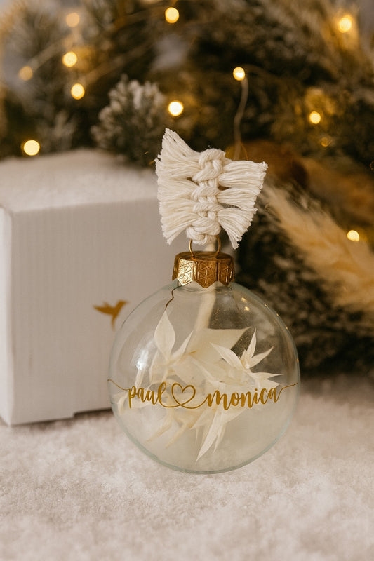 Personalisierte Weihnachtskugel aus Glas mit goldenem Schriftzug ‚paul ♥ monica‘, gefüllt mit weißen Trockenblumen und handgemachtem Makramee-Aufhänger, dekoriert vor einem weihnachtlichen Hintergrund mit Lichterkette und Tannenzweigen.