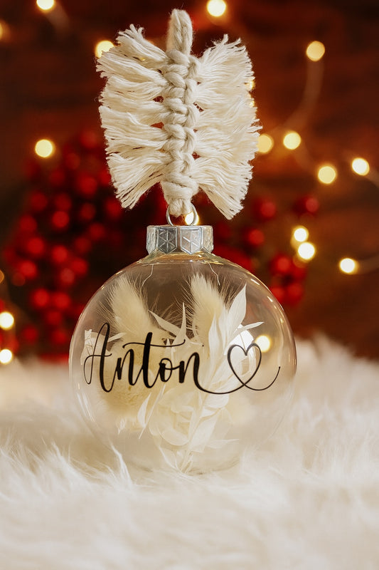 Personalisierte Weihnachtskugel aus Glas mit silbernem Kranz und handgemachtem Makramee-Aufhänger, gefüllt mit weißen Trockenblumen. Namensaufdruck ‚Anton‘ in schwarzer Schrift mit Herzsymbol, dekoriert vor weihnachtlicher Lichterkulisse.