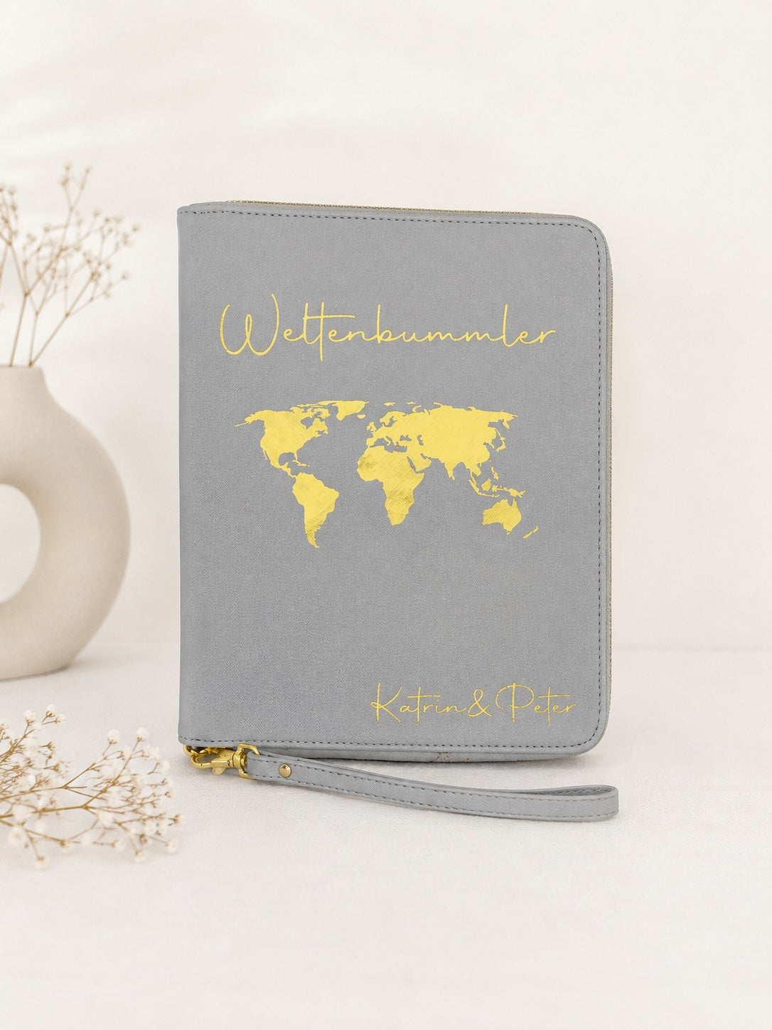 Grauer personalisierter Reiseorganizer mit goldenem Schriftzug „Weltenbummler“, Weltkarte und den Namen Katrin und Peter vor hellem neutralem Hintergrund.