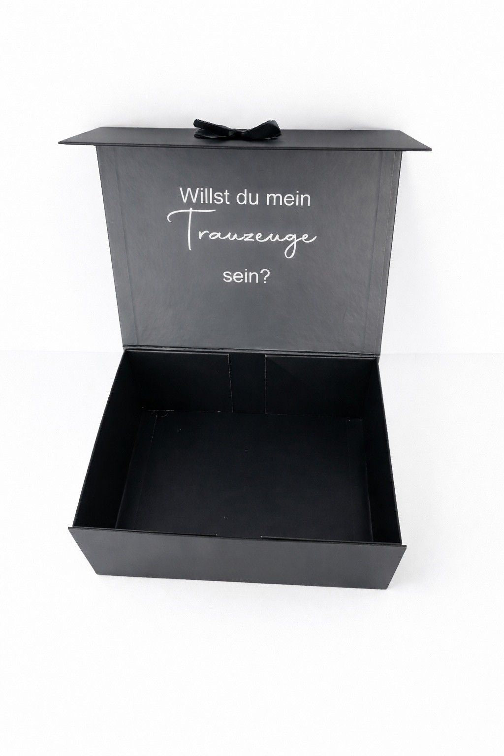 Offene schwarze Geschenkbox mit der Aufschrift ‚Willst du mein Trauzeuge sein?‘ auf hellem, minimalistischem Hintergrund.