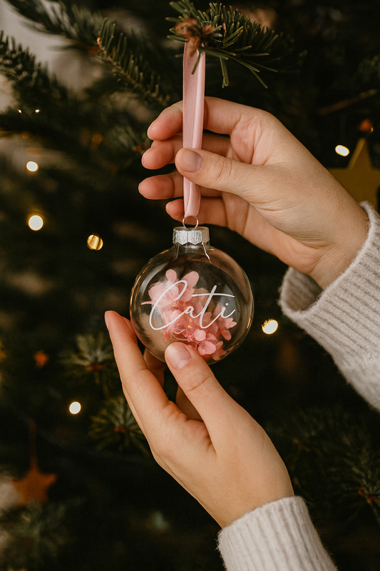 Hand hält personalisierte Weihnachtskugel aus Glas mit Namen Cati, gefüllt mit rosa Trockenblumen und rosa Band, hängend am Weihnachtsbaum.