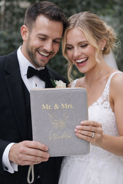 Glückliches Brautpaar betrachtet gemeinsam einen personalisierten Dokumentenorganizer mit der Aufschrift „Mr. & Mrs.“ als Hochzeitsgeschenk.