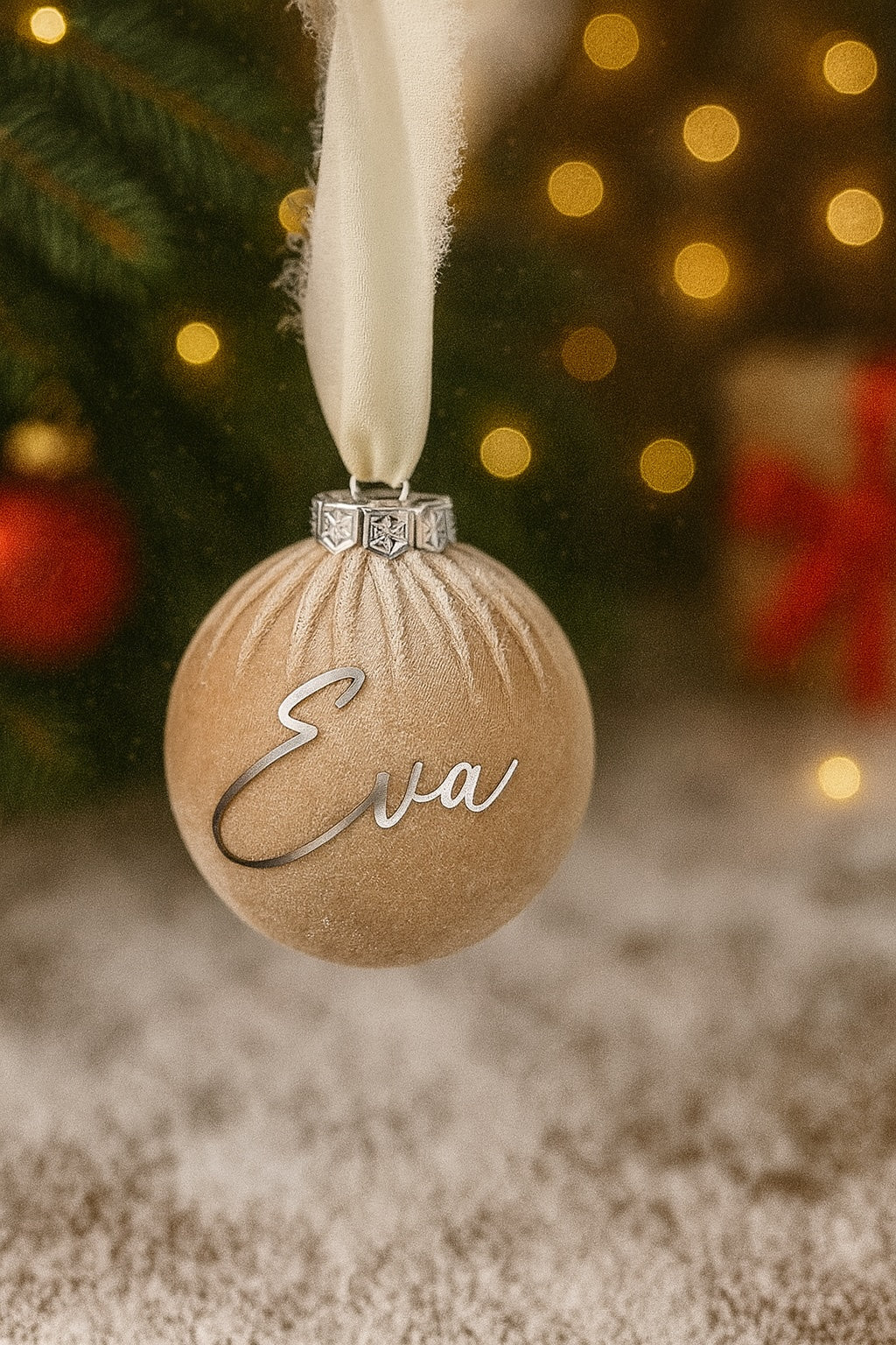 Personalisierte Weihnachtskugel in Beige, Ø 8 cm, beflockt mit samtiger Oberfläche und silbernem Kranz, verziert mit dem Namen ‚Eva‘ in eleganter Schrift. Aufgehängt am weißen Chiffonband, vor weihnachtlichem Hintergrund mit Tannenbaum, Lichtern und Geschenken.