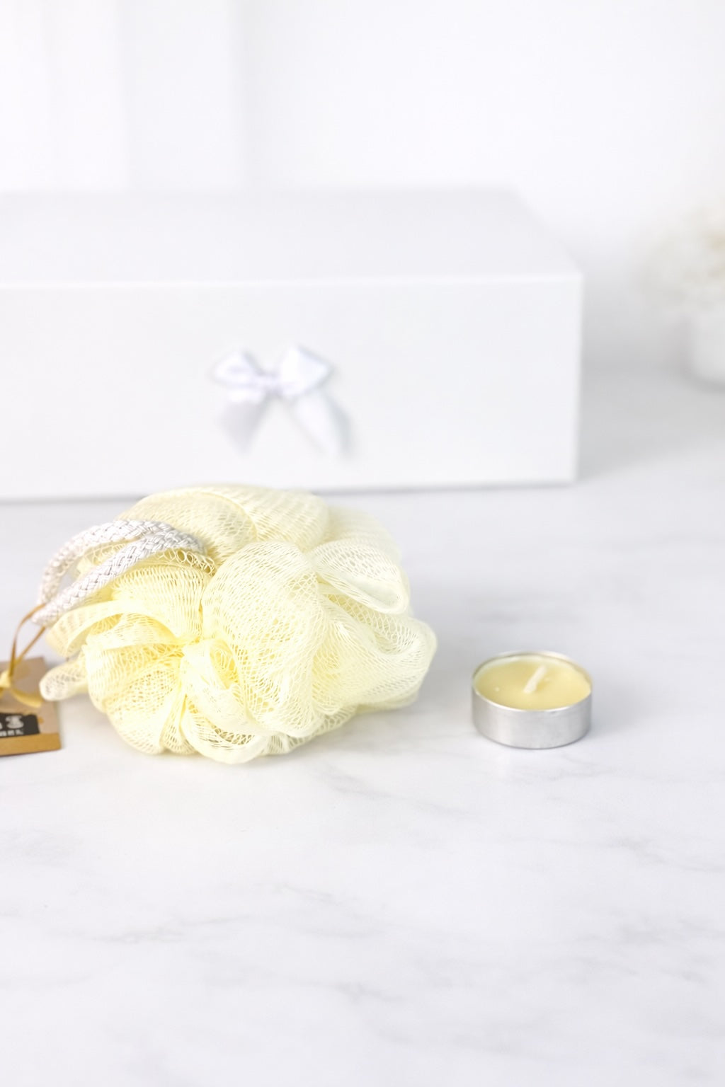 Duschschwamm und Teelicht als Bade-Accessoires auf hellem Hintergrund mit Geschenkbox im Hintergrund