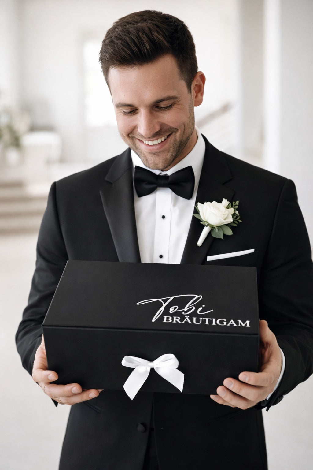 Glücklicher Bräutigam im schwarzen Anzug hält eine personalisierte schwarze Geschenkbox mit Aufdruck „Tobi Bräutigam“ und weißer Schleife in den Händen.