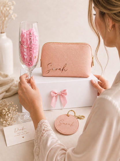 Lifestyle-Foto einer personalisierten Brautjungfer-Geschenkbox mit Kosmetiktasche, Sektglas und Anhänger mit dem Namen Sarah, während eine Frau im Satinmorgenmantel das Geschenk betrachtet, ohne dass ihr Gesicht zu sehen ist.