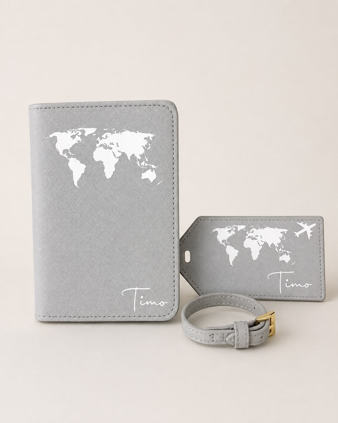 Personalisierte graue Reisepasshülle mit passendem Kofferanhänger und Gepäckriemen, verziert mit weißem Weltkartenmotiv und Namen „Timo“. Hochwertiges Reiseaccessoire-Set auf neutralem Hintergrund.
