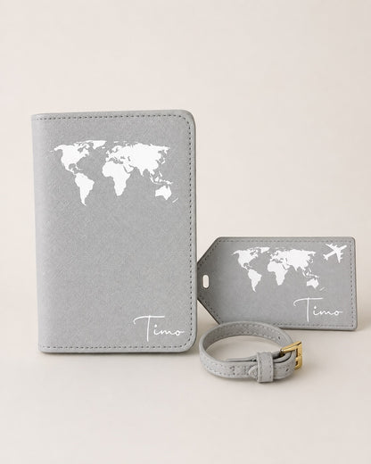 Personalisierte graue Reisepasshülle mit passendem Kofferanhänger und Gepäckriemen, verziert mit weißem Weltkartenmotiv und Namen „Timo“. Hochwertiges Reiseaccessoire-Set auf neutralem Hintergrund.