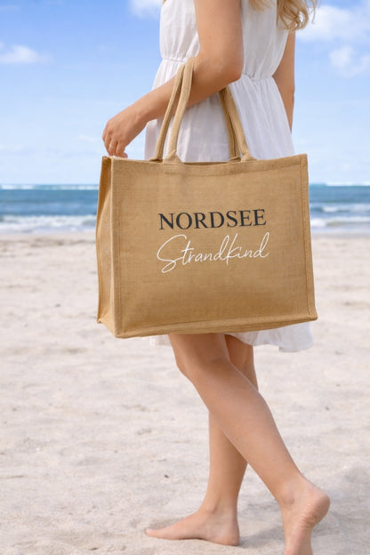 Frau trägt Jute Strandtasche „Nordsee Strandkind“ aus Naturjute am Strand mit Meer im Hintergrund
