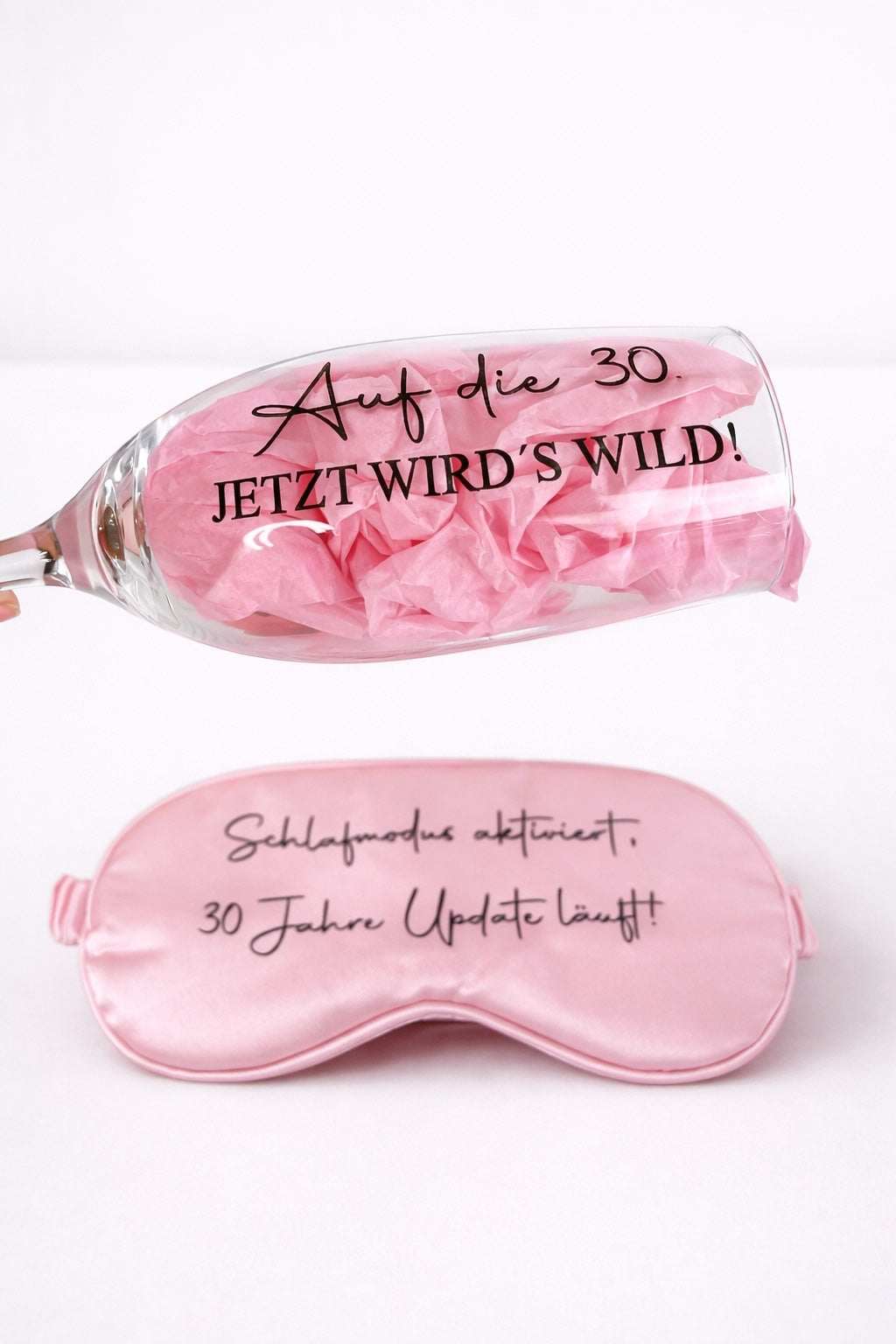 Sektglas mit Aufdruck ‚Auf die 30 – jetzt wird’s wild!‘ und rosa Schlafmaske als Geschenkset zum 30. Geburtstag vor hellem Hintergrund