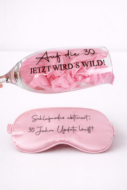 Sektglas mit Aufdruck ‚Auf die 30 – jetzt wird’s wild!‘ und rosa Schlafmaske als Geschenkset zum 30. Geburtstag vor hellem Hintergrund
