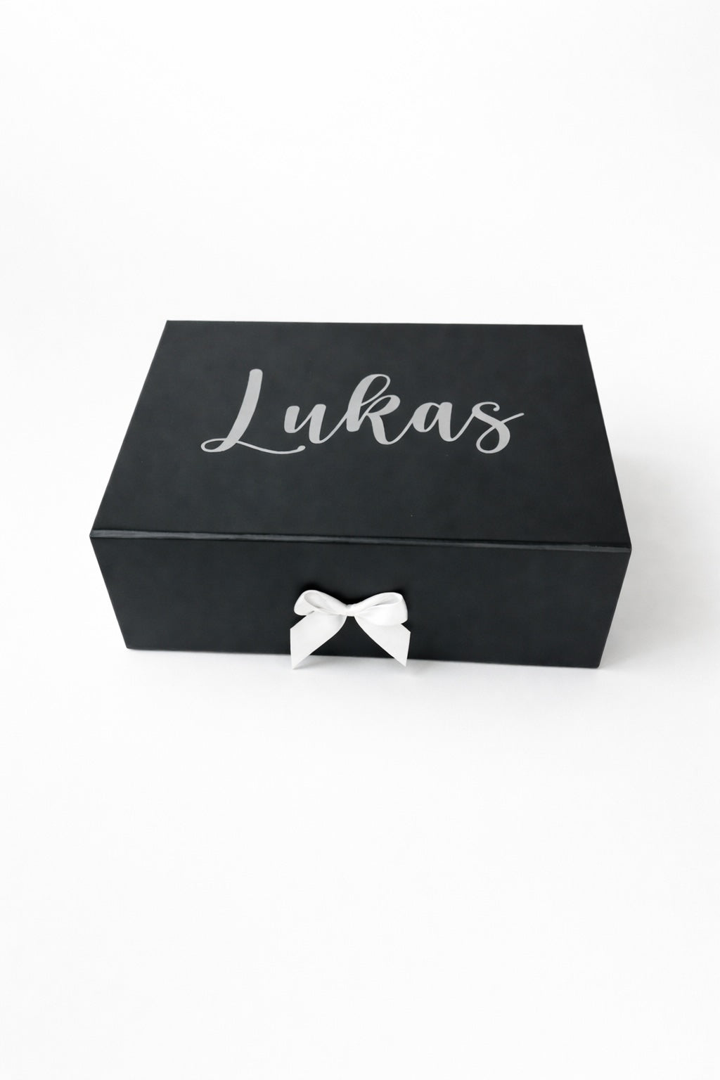 Schwarze, personalisierte Geschenkbox mit dem Namen Lukas in silberner Schrift und weißer Schleife auf hellem Hintergrund.