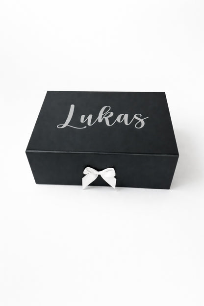 Schwarze, personalisierte Geschenkbox mit dem Namen Lukas in silberner Schrift und weißer Schleife auf hellem Hintergrund.