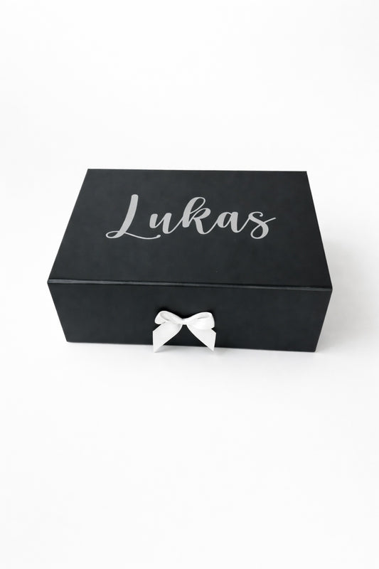 Schwarze, personalisierte Geschenkbox mit dem Namen Lukas in silberner Schrift und weißer Schleife auf hellem Hintergrund.