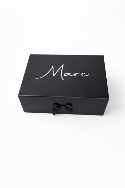 Schwarze, personalisierte Geschenkbox mit dem Namen Marc in weißer Schrift und schwarzer Schleife auf hellem, minimalistischem Hintergrund.“