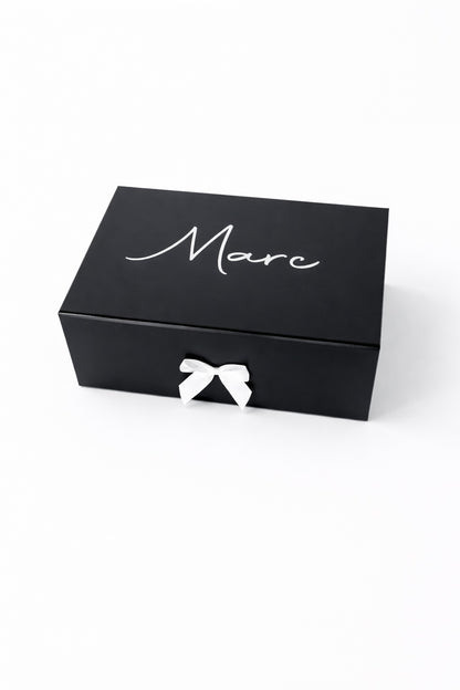 Schwarze, personalisierte Geschenkbox mit dem Namen Marc in weißer Schrift und weißer Schleife auf hellem, minimalistischem Hintergrund.