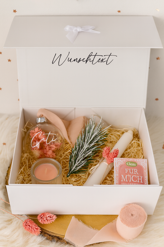 Offene weiße Geschenkbox mit personalisierbarer Aufschrift ‚Wunschtext‘ im Deckel. In der Box liegen eine durchsichtige Christbaumkugel mit dem Namen ‚Cati‘, eine rosa Kerze im Glas, ein Stück Seife mit der Aufschrift ‚Für mich‘, eine weiße Kerze mit rosa Trockenblume und ein dekorierter Tannenzweig. Der Hintergrund ist weihnachtlich mit Weihnachtssternen