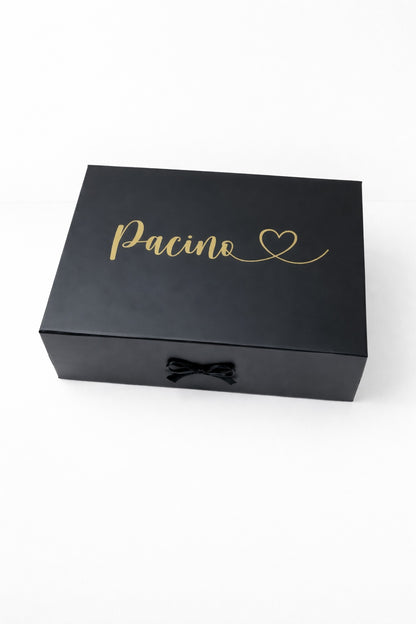 Schwarze Geschenkbox mit goldenem Schriftzug ‚Pacino‘ und Herz, mit schwarzer Schleife auf hellem, minimalistischem Hintergrund