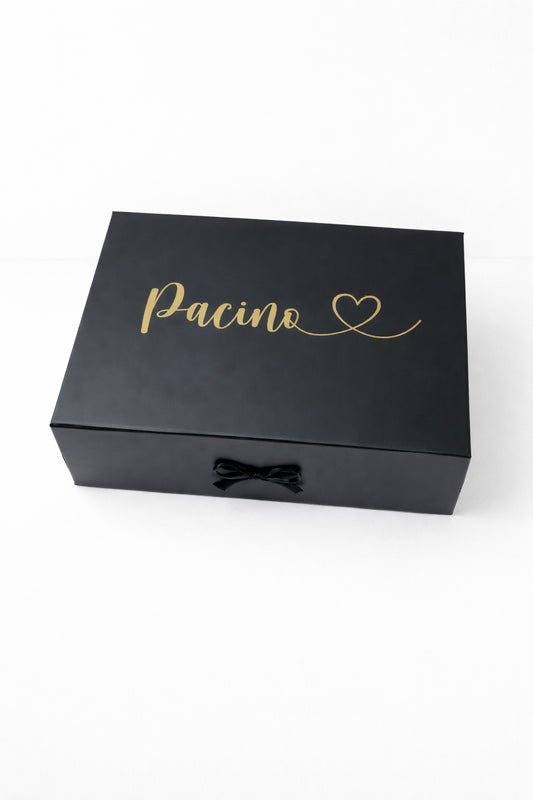 Schwarze Geschenkbox mit goldenem Schriftzug ‚Pacino‘ und Herz, mit schwarzer Schleife auf hellem, minimalistischem Hintergrund