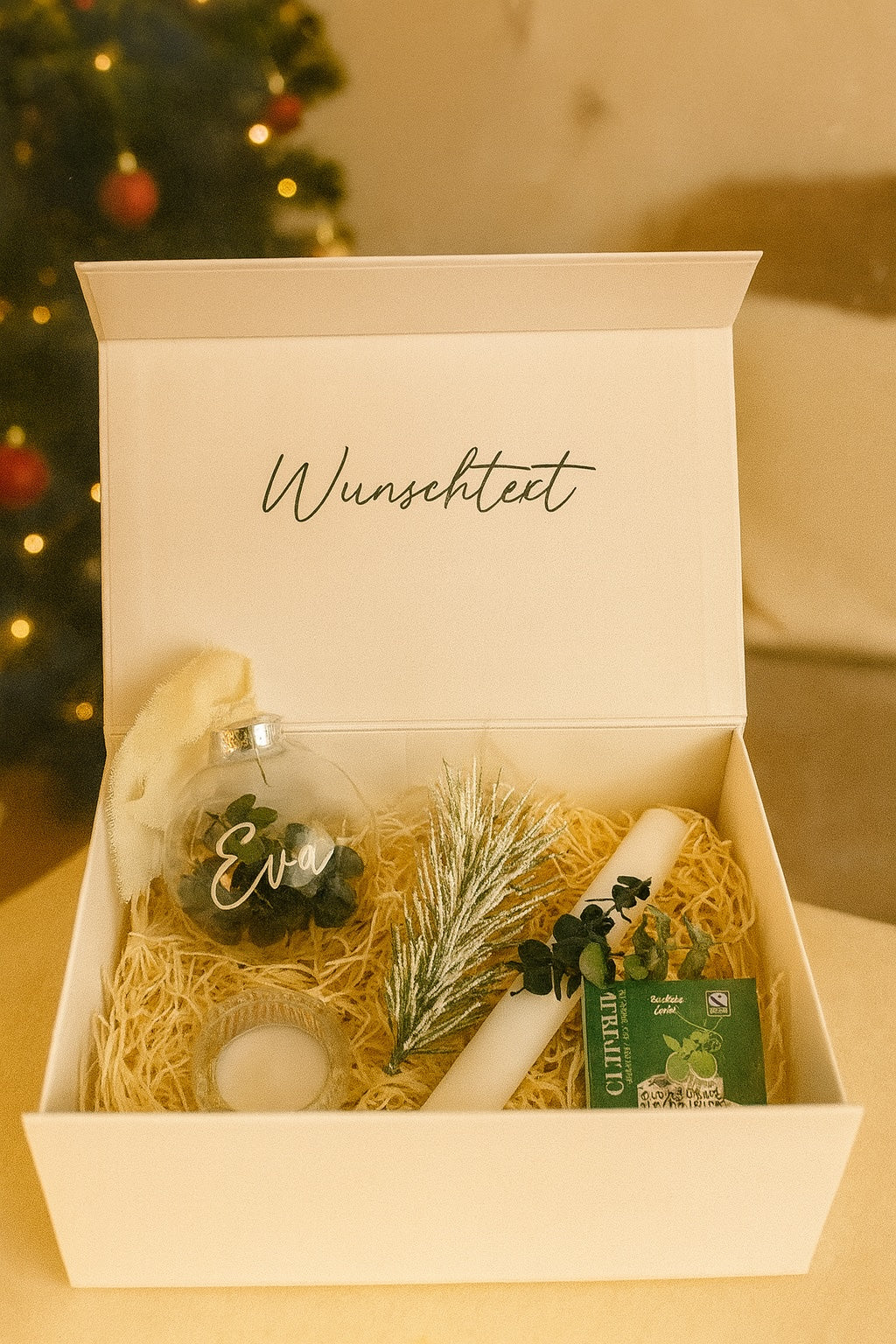 Weiße Geschenkbox mit der Aufschrift ‚Wunschtext‘, gefüllt mit einer personalisierten Weihnachtskugel mit Namen Eva, einer weißen Kerze mit Eukalyptus, einer kleinen Tannenzweig-Deko, einem Teelichthalter und einer Packung Cuppa Tee. Die Box ist weihnachtlich dekoriert und offen präsentiert.