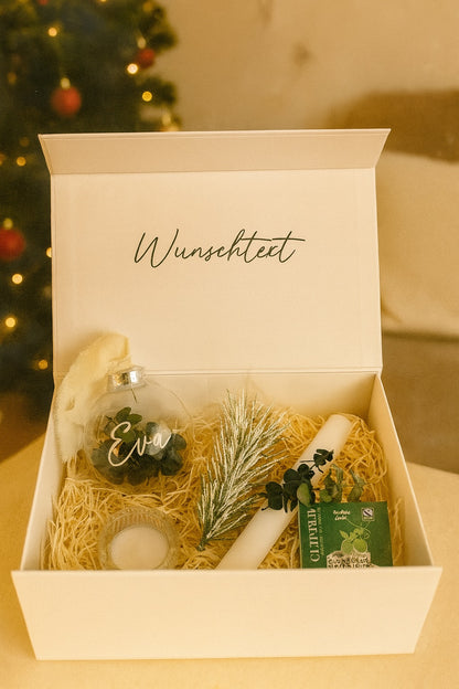 Weiße Geschenkbox mit der Aufschrift ‚Wunschtext‘, gefüllt mit einer personalisierten Weihnachtskugel mit Namen Eva, einer weißen Kerze mit Eukalyptus, einer kleinen Tannenzweig-Deko, einem Teelichthalter und einer Packung Cuppa Tee. Die Box ist weihnachtlich dekoriert und offen präsentiert.