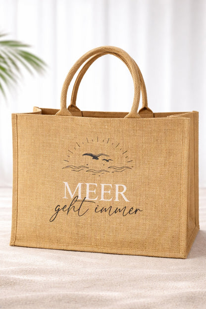Natürliche Jute Strandtasche mit Aufdruck „Meer geht immer“, große Beach Bag aus Jute mit stabilen Griffen vor hellem neutralem Hintergrund.