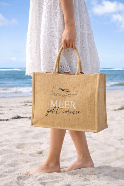 Frau trägt eine Jute Strandtasche mit Aufdruck „Meer geht immer“ am Strand, natürliche Beach Bag aus Jute mit Meer und Sand im Hintergrund.