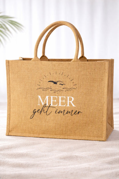 Jute Strandtasche mit Aufdruck „Meer geht immer“ in weißer Schrift vor hellem neutralem Hintergrund, robuste Naturtasche mit stabilen Griffen im maritimen Design.
