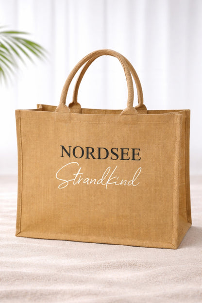 Jute Strandtasche „Nordsee Strandkind“ mit schwarzem und weißem Aufdruck auf naturfarbener Tasche vor hellem neutralem Hintergrund.
