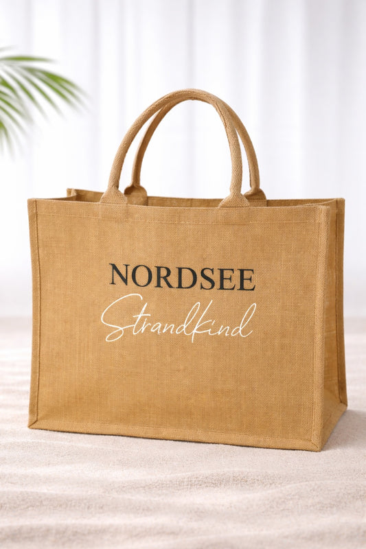 Jute Strandtasche „Nordsee Strandkind“ mit schwarzem und weißem Aufdruck auf naturfarbener Tasche vor hellem neutralem Hintergrund.

