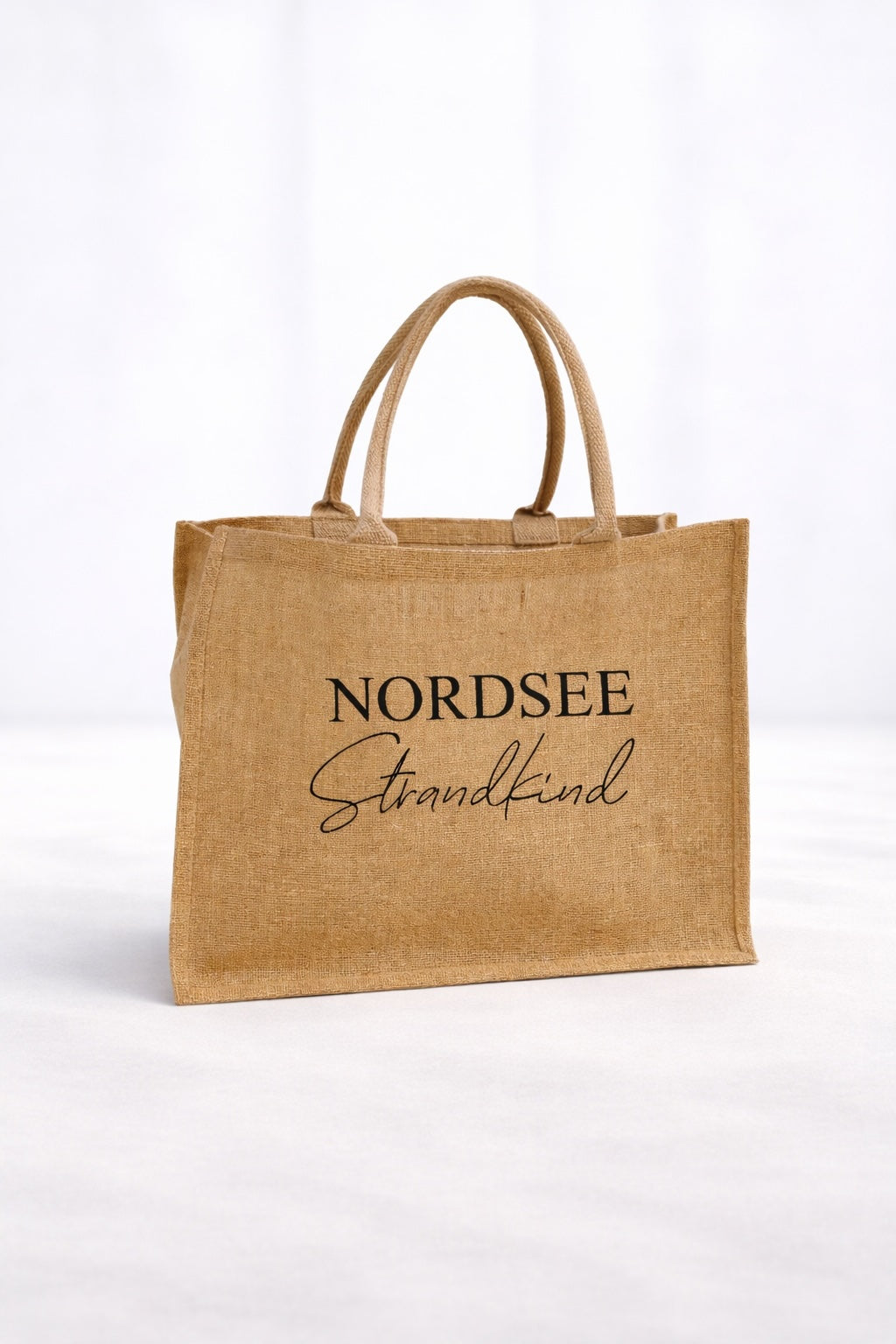 Naturfarbene Jute Strandtasche „Nordsee Strandkind“ mit schwarzem Aufdruck vor hellem neutralem Hintergrund.