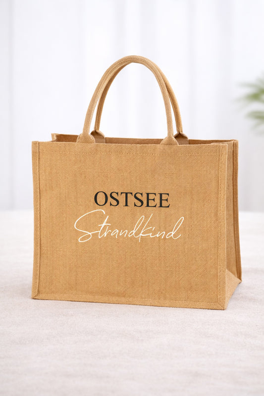 Jute Strandtasche in Naturfarbe mit Aufdruck „OSTSEE Strandkind“ freigestellt vor hellem, neutralem Hintergrund.