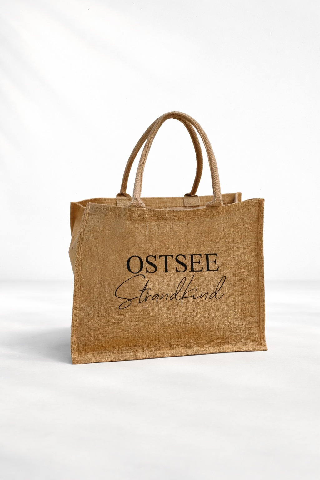 Jute Strandtasche „Ostsee Strandkind“ mit schwarzem Aufdruck auf naturfarbener Tasche vor hellem neutralem Hintergrund