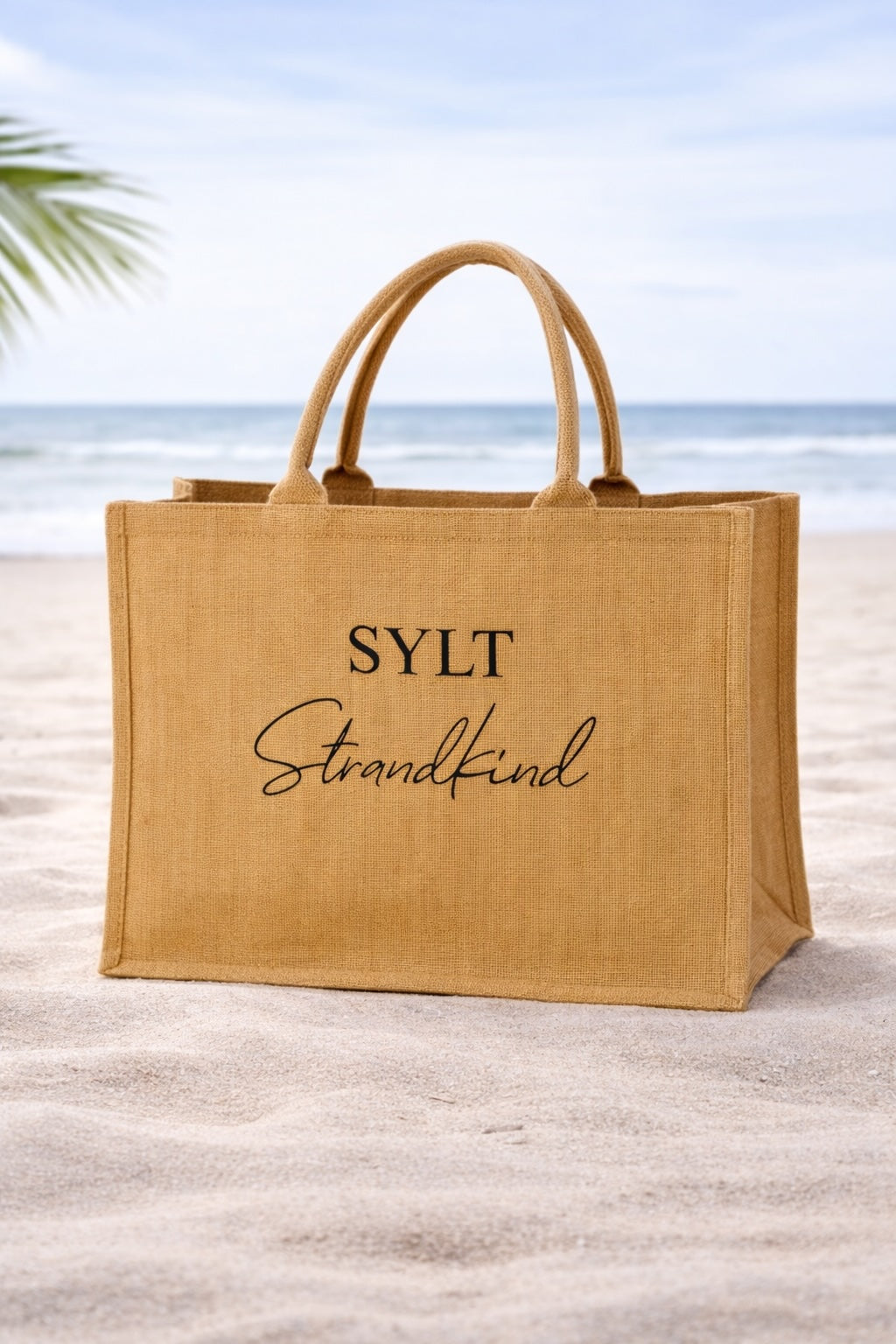 Jute Strandtasche mit schwarzem Aufdruck „SYLT Strandkind“ steht im Sand vor Meer und blauem Himmel am Strand.