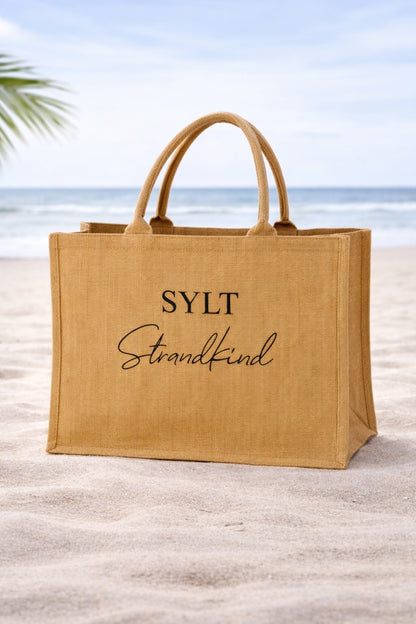 Jute Strandtasche mit schwarzem Aufdruck „SYLT Strandkind“ steht im Sand vor Meer und blauem Himmel am Strand.