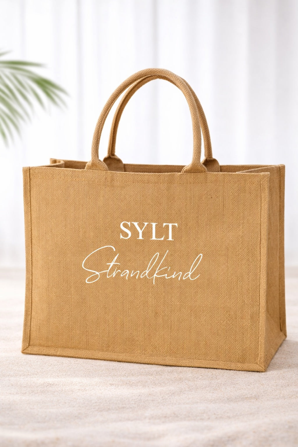 Große Jute-Strandtasche in Naturfarbe mit weißem Aufdruck „SYLT Strandkind“, freigestellt vor hellem, neutralem Hintergrund.