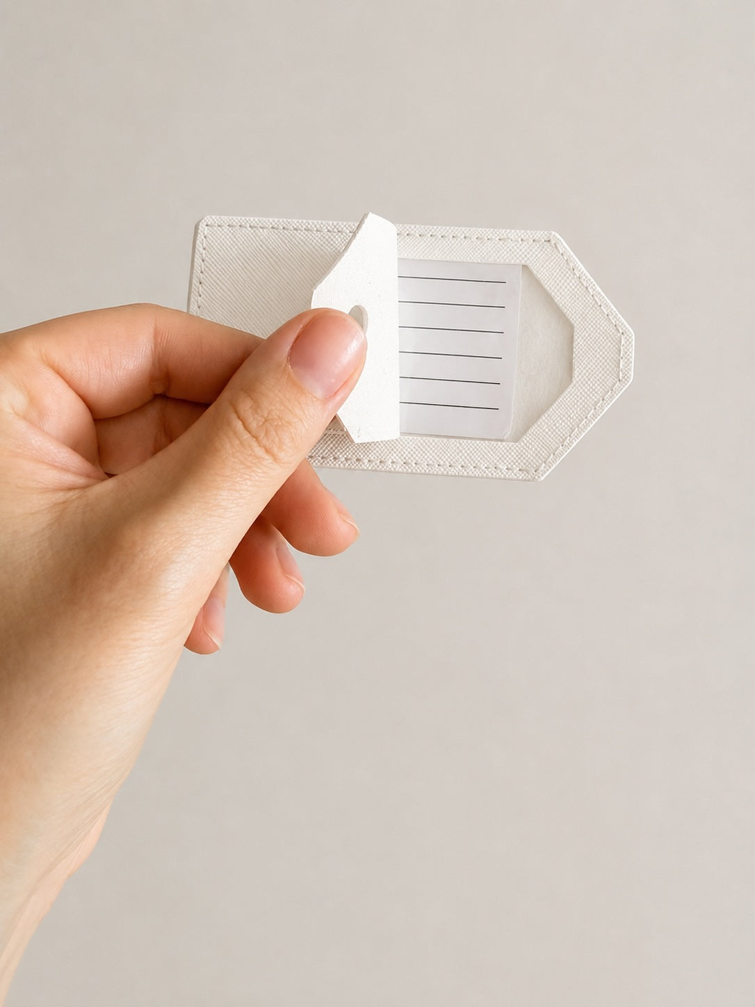 Hand hält einen weißen Kofferanhänger mit geöffnetem Adressfeld auf schlichtem, neutralem Hintergrund, Nahaufnahme eines Reiseaccessoires.
