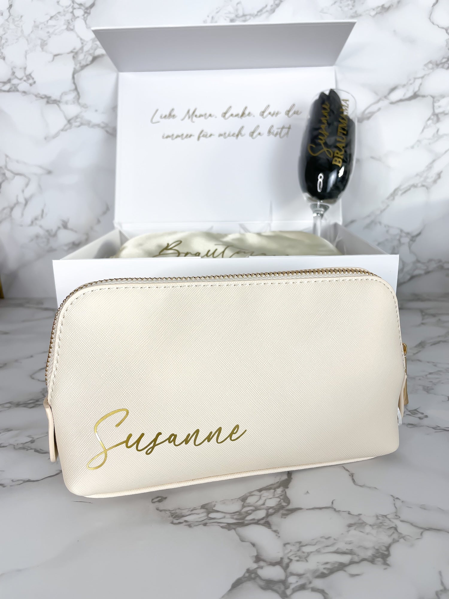 Personalisierte Kosmetiktasche mit Namen als Teil einer Geschenkbox für die Brautmama, arrangiert mit Schlafmaske und Sektglas in geöffneter Box.