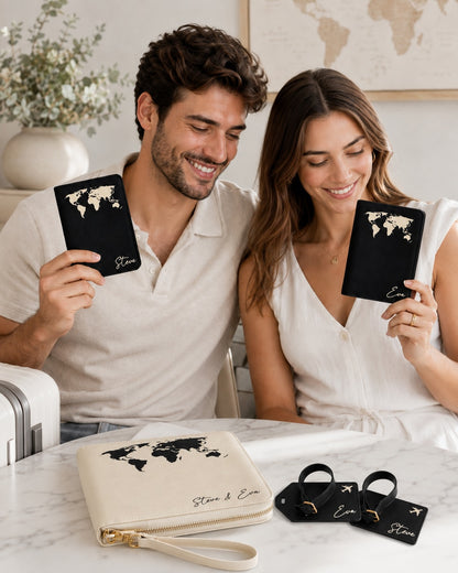 Lifestyle-Foto eines lächelnden Paares an einem Tisch. Beide halten schwarze personalisierte Reisepasshüllen mit Weltkarten-Design und den Namen „Steve“ und „Eva“. Auf dem Tisch liegen eine beige Reise-Organizer-Tasche sowie schwarze Gepäckanhänger mit Flugzeugsymbol. Die Szene wirkt hell, warm und reisefreudig in einem gemütlichen Wohnambiente.