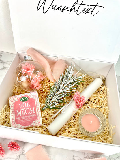 Personalisierte weiße Geschenkbox mit Wunschtext, daneben der Inhalt: Glas-Weihnachtskugel mit rosa Trockenblumen und Chiffonband, weißer Kunst-Tannenzweig mit Schnee-Effekt, weiße Stabkerze mit rosa Trockenblumen, Kräutertee von Cleo’s und rosa Duft-Teelicht im Glas.