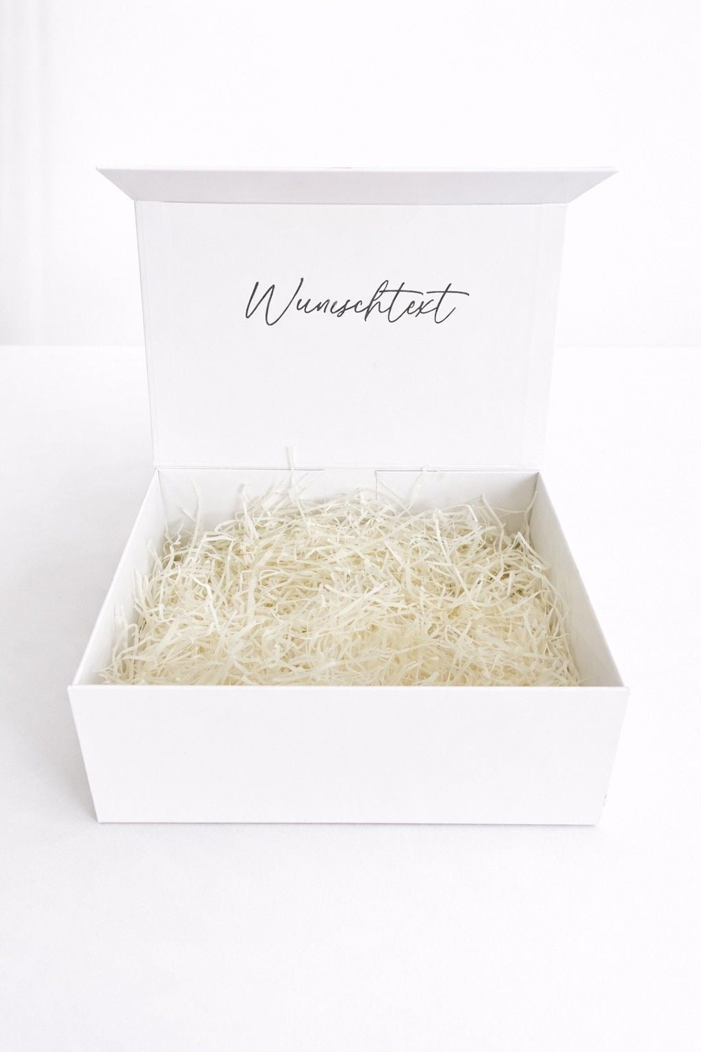 Offene weiße Geschenkbox mit Wunschtext auf dem Deckel und Füllmaterial, fotografiert vor hellem, minimalistischem Hintergrund