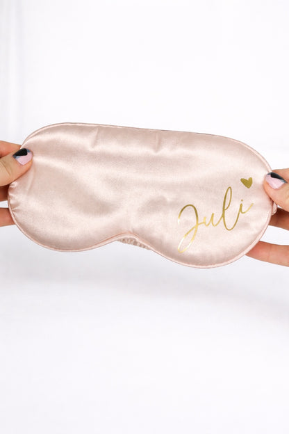Personalisierte Schlafmaske aus Satin in Rosa mit Name ‚Juli‘ in Gold, gehalten vor hellem, minimalistischem Hintergrund