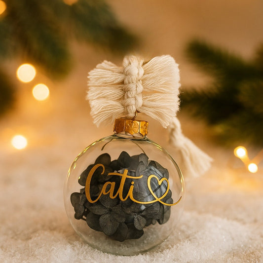 Personalisierte Weihnachtskugel aus Glas mit goldenem Namensaufdruck ‚Cati‘ und Herzsymbol, gefüllt mit dunklen Eukalyptus-Trockenblättern, goldenem Kugelkranz und handgemachtem Makramee-Aufhänger, dekoriert vor weihnachtlicher Lichterkulisse.